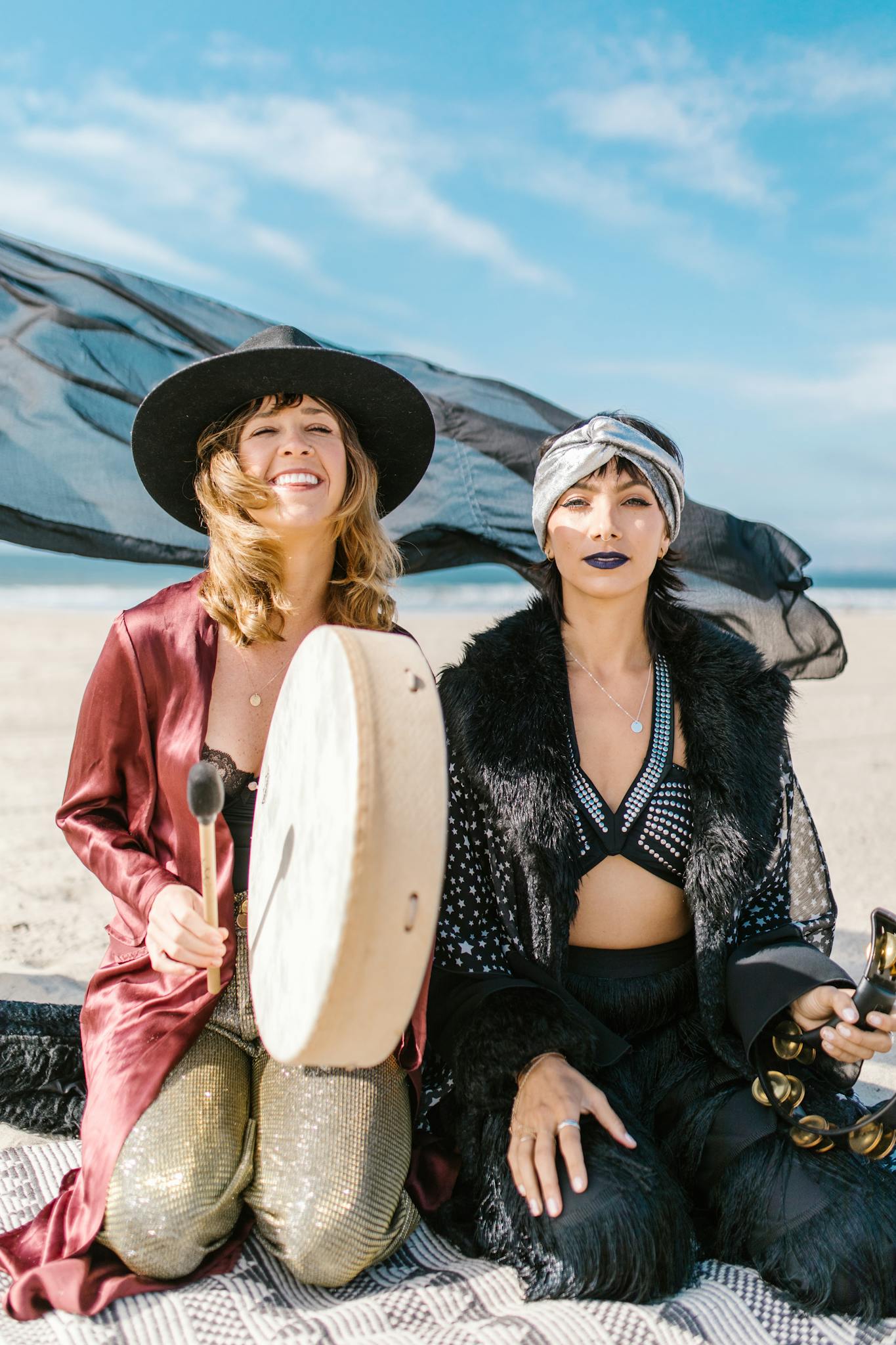 Two women in bohemian attire with drums and tambourine on a sunny beach.